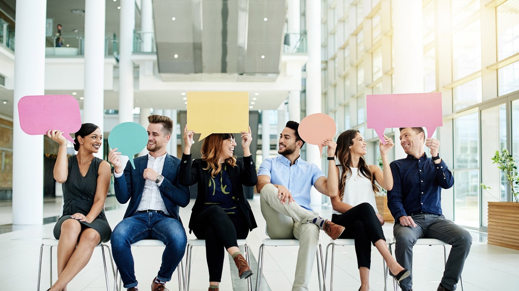 Heterogene Personen aus einem Team sitzen auf Stühlen und halten Sprechblasen aus Pappe in die Luft